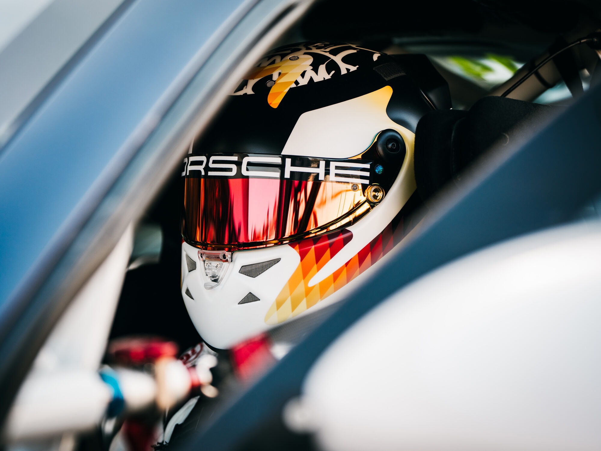 Rennfahrerhelm mit Porsche-Schriftzug und verspiegeltem Visier im Cockpit, gesehen durch das offene Seitenfenster