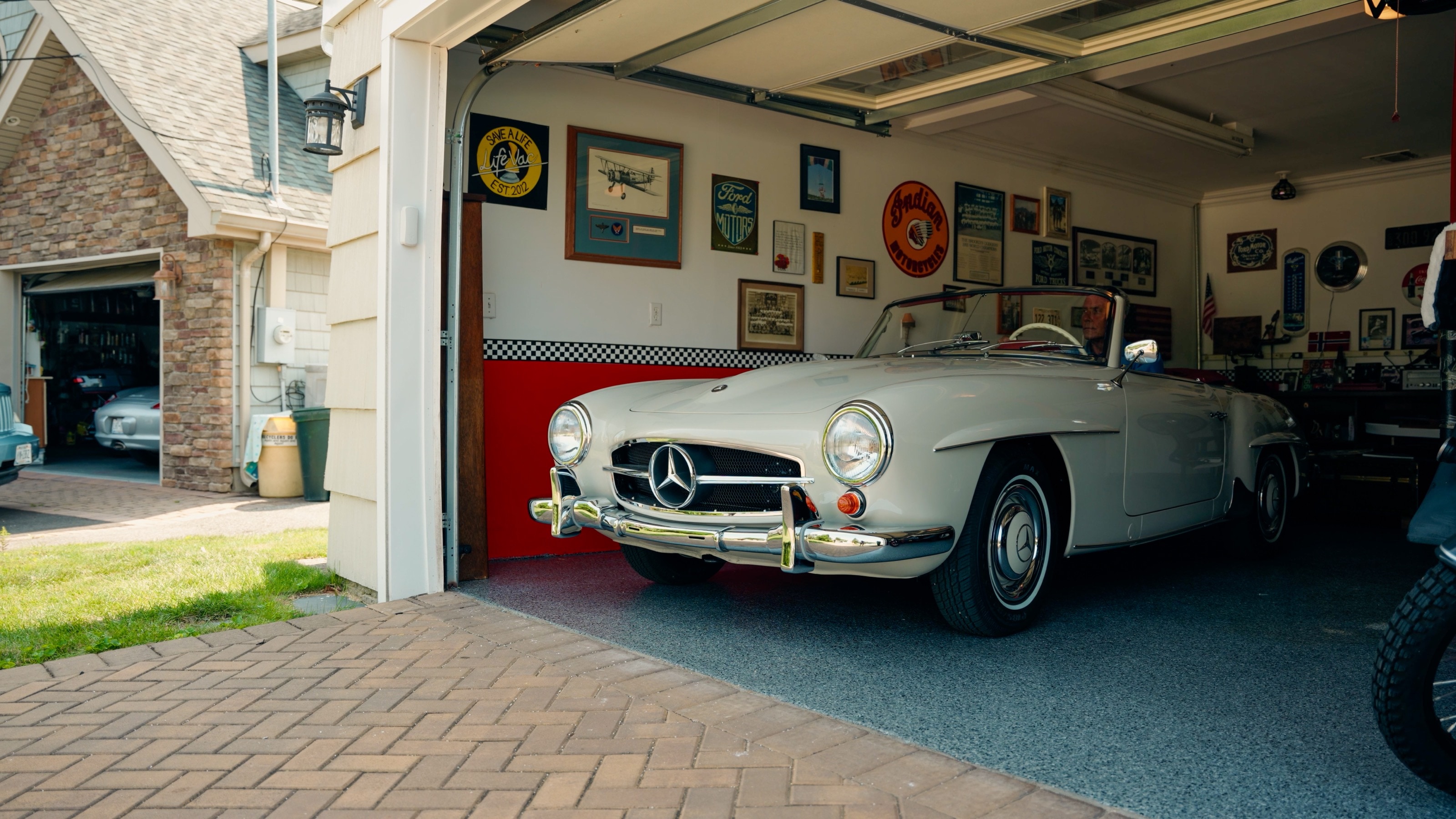 Der cremeweiße 190 SL steht in einer offenen Garage mit roter Wand und Erinnerungsstücken