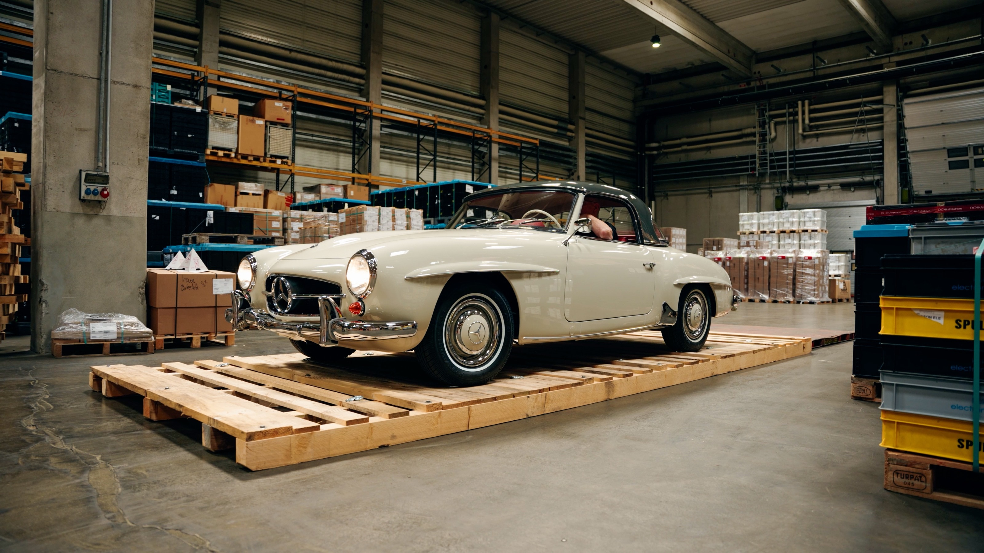 Der 190 SL steht auf Holzpaletten in einer Lagerhalle zwischen Regalen und Kisten