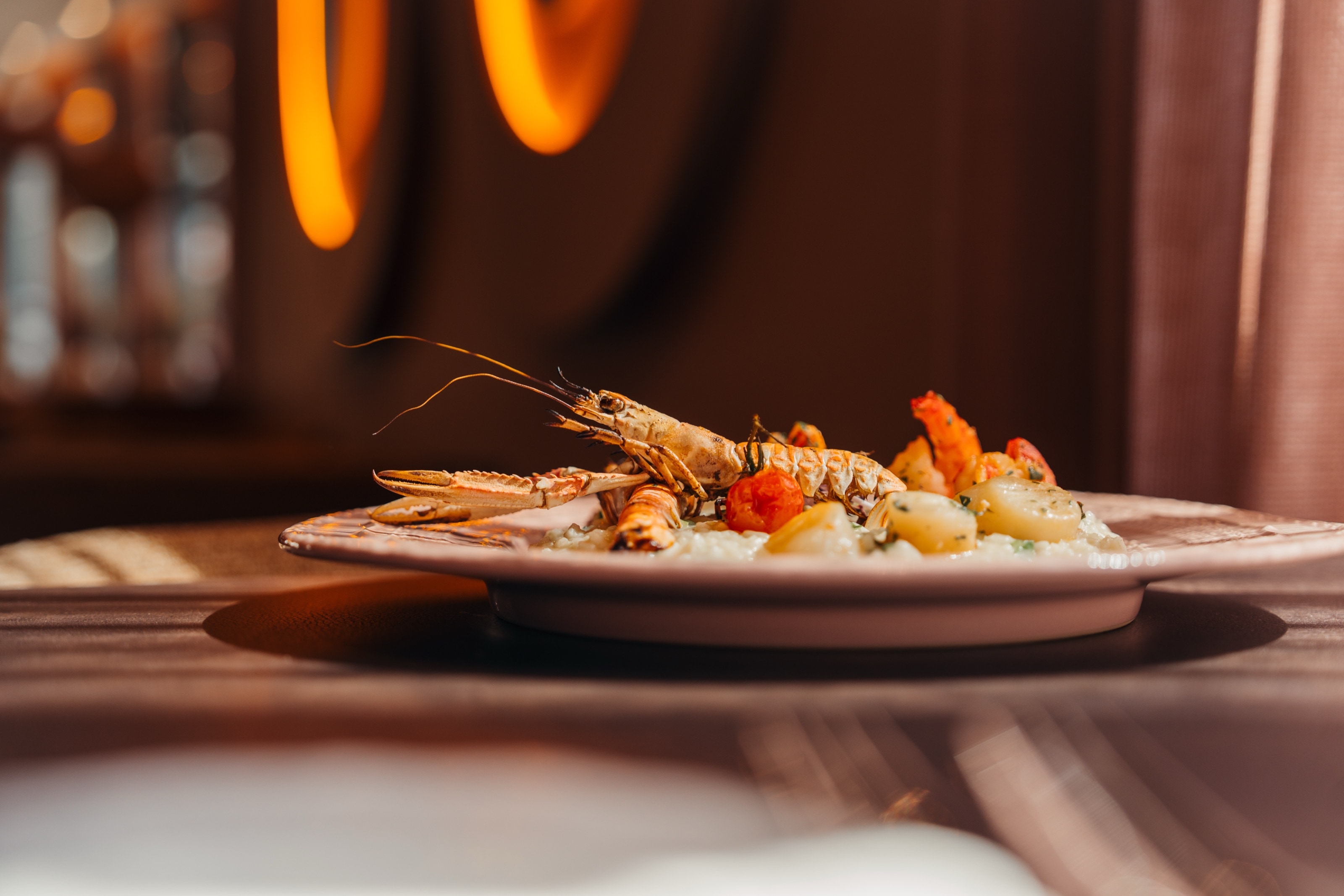 Langustine auf Risotto mit Garnelen und Kirschtomaten, auf Tellerhöhe fotografiert, dahinter warmes Licht im Gastraum