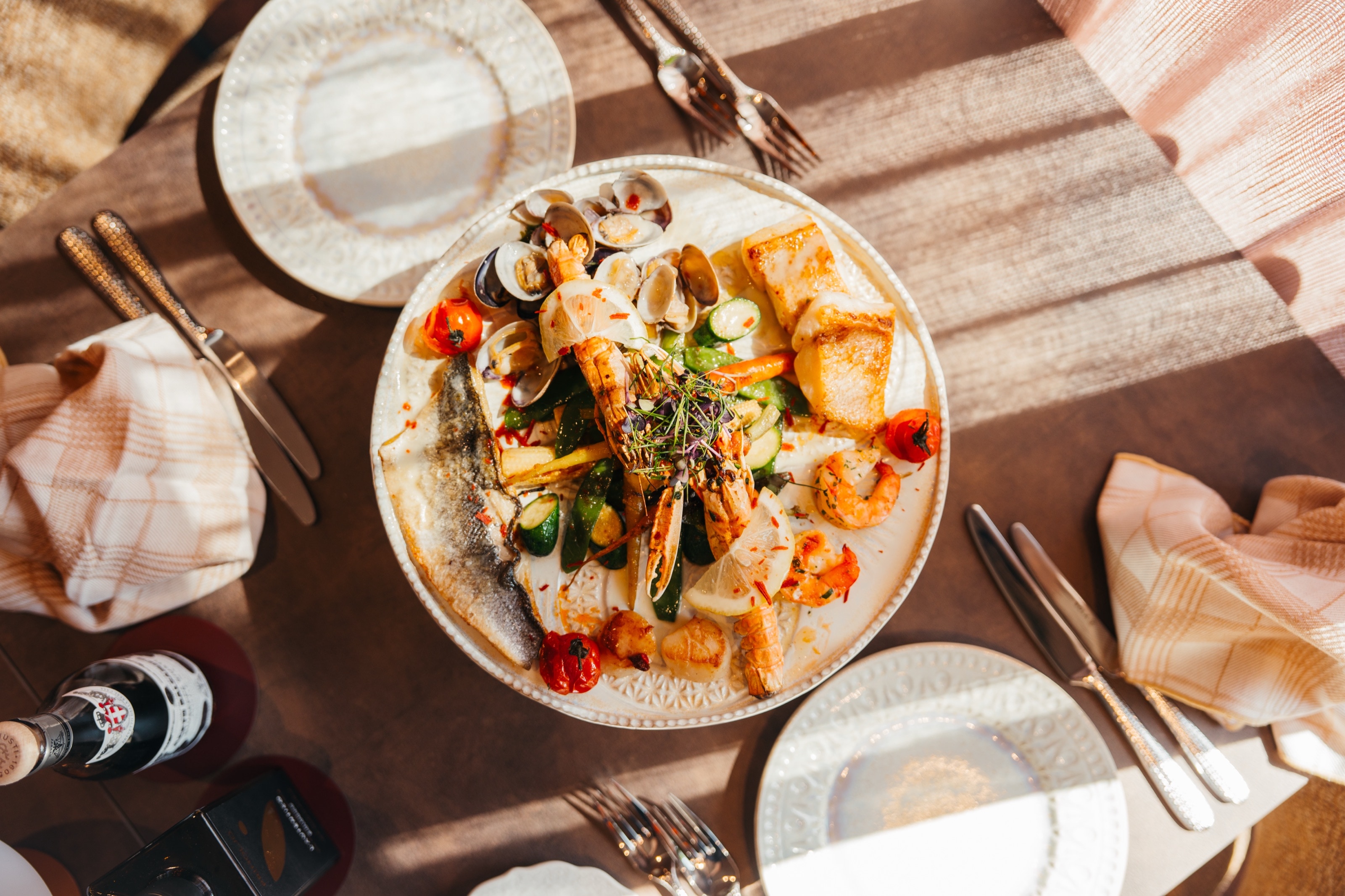 Meeresfrüchteplatte von oben mit Fischfilet, Venusmuscheln, Garnelen und gegrilltem Gemüse, Sonnenstreifen auf dem Tisch