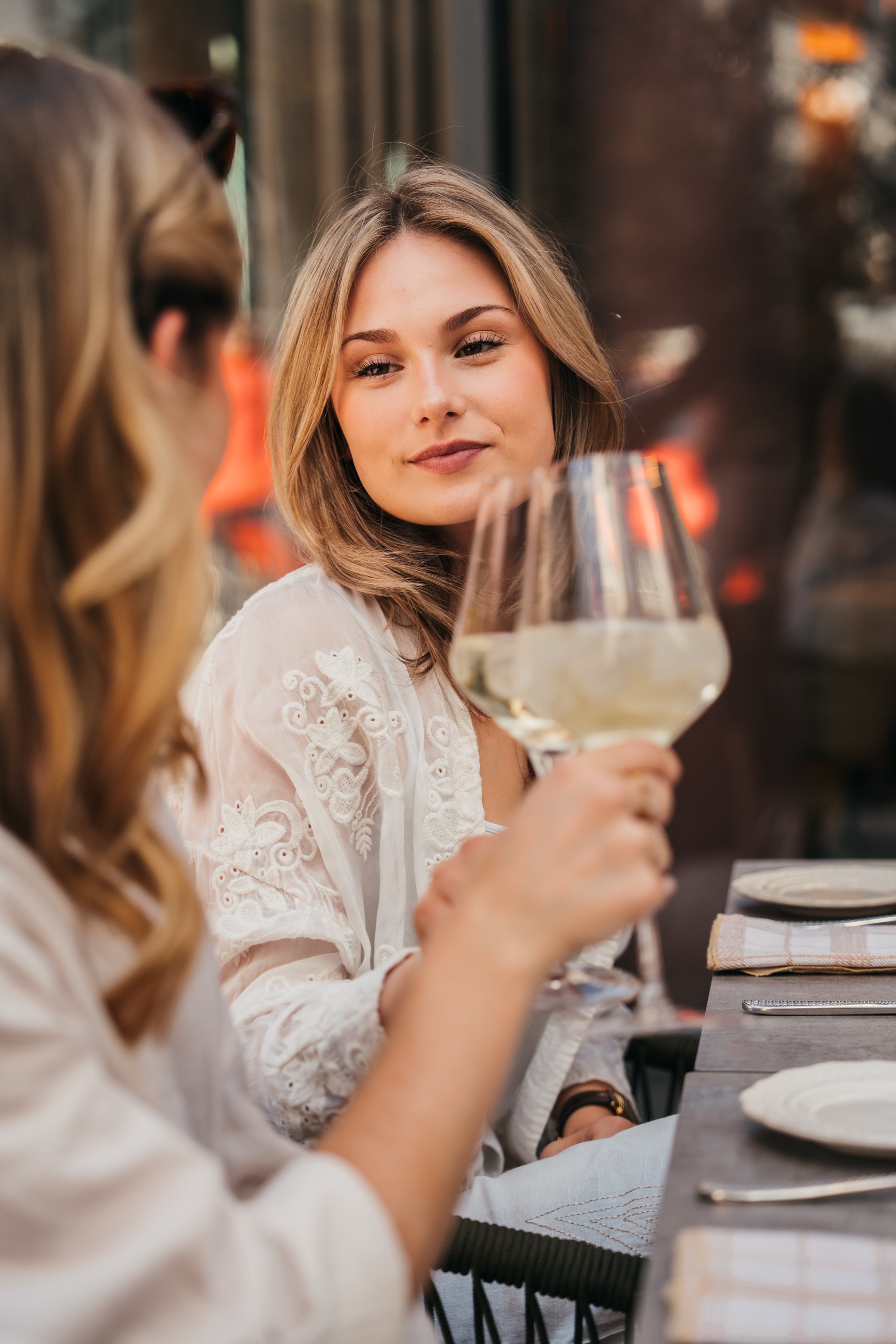 Zwei Frauen am Tisch, eine hebt ihr Weißweinglas, die andere im weißen, bestickten Oberteil lächelt ihr zu