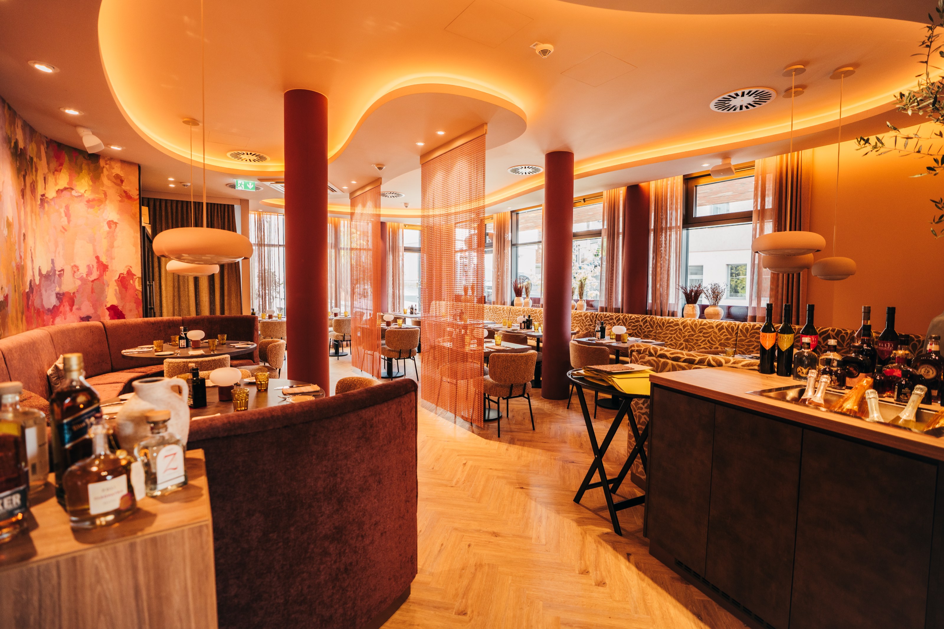 Weiter Blick durch das Restaurant mit weinroten Säulen, geschwungener Lichtdecke und Polsternischen, im Vordergrund die Bar