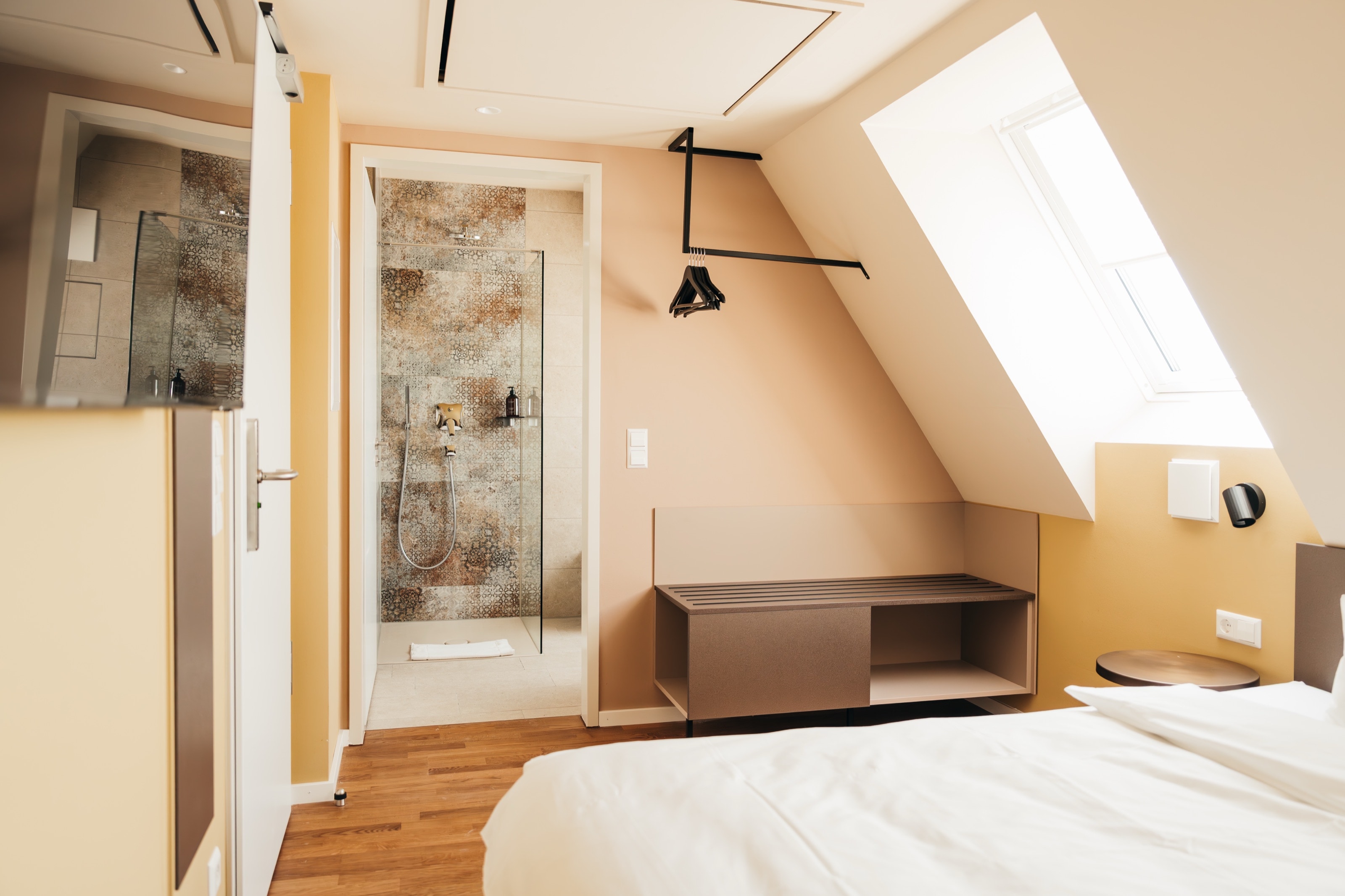 Zimmer unter der Dachschräge mit Dachfenster, offener Kleiderstange und Blick in die Dusche mit ornamental gemusterten Fliesen