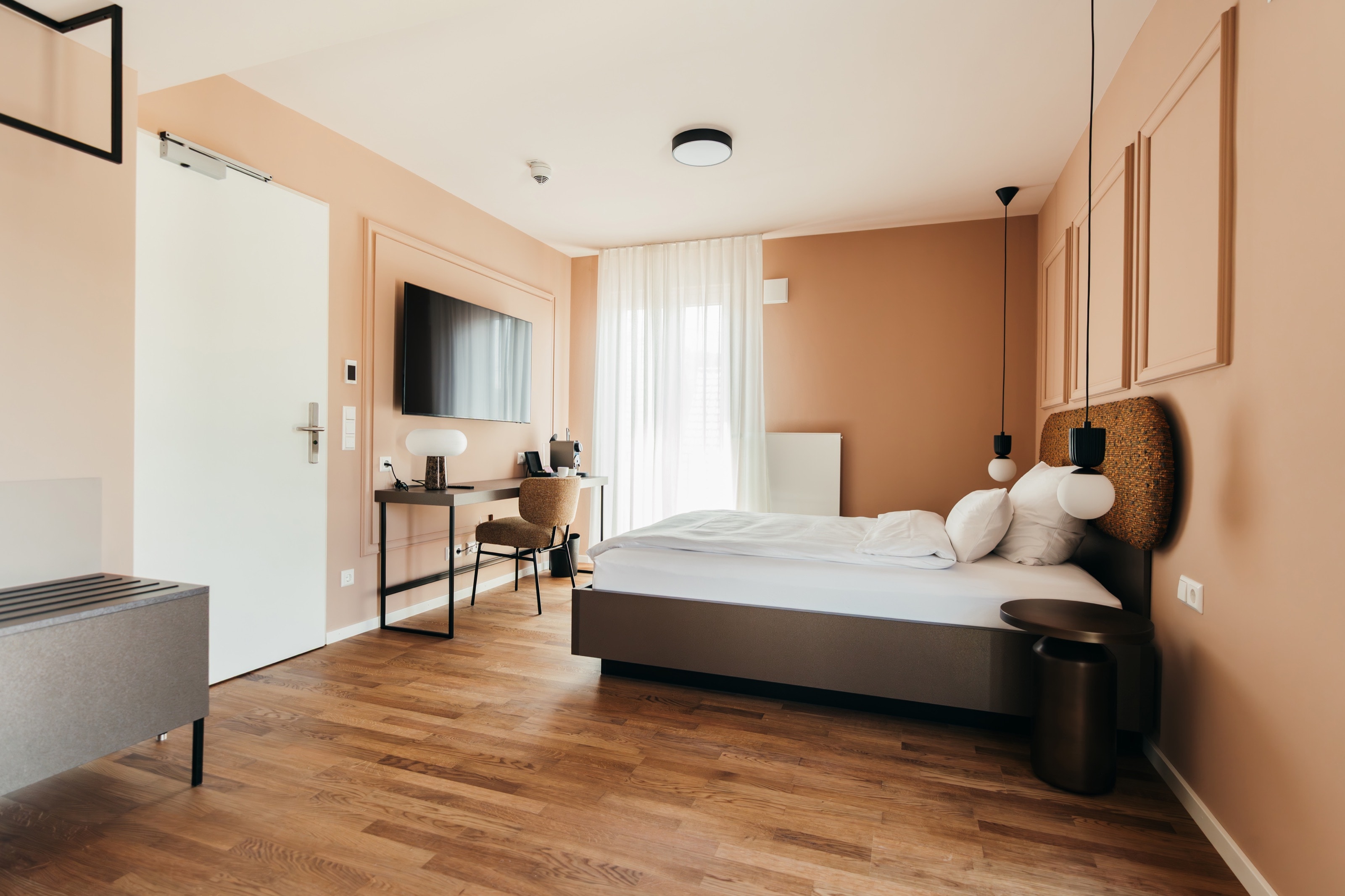 Suite mit pfirsichfarbenen Wänden und profilierten Wandfeldern, Doppelbett mit strukturiertem Kopfteil und Kugel-Pendelleuchten, links Schreibtisch und Fernseher