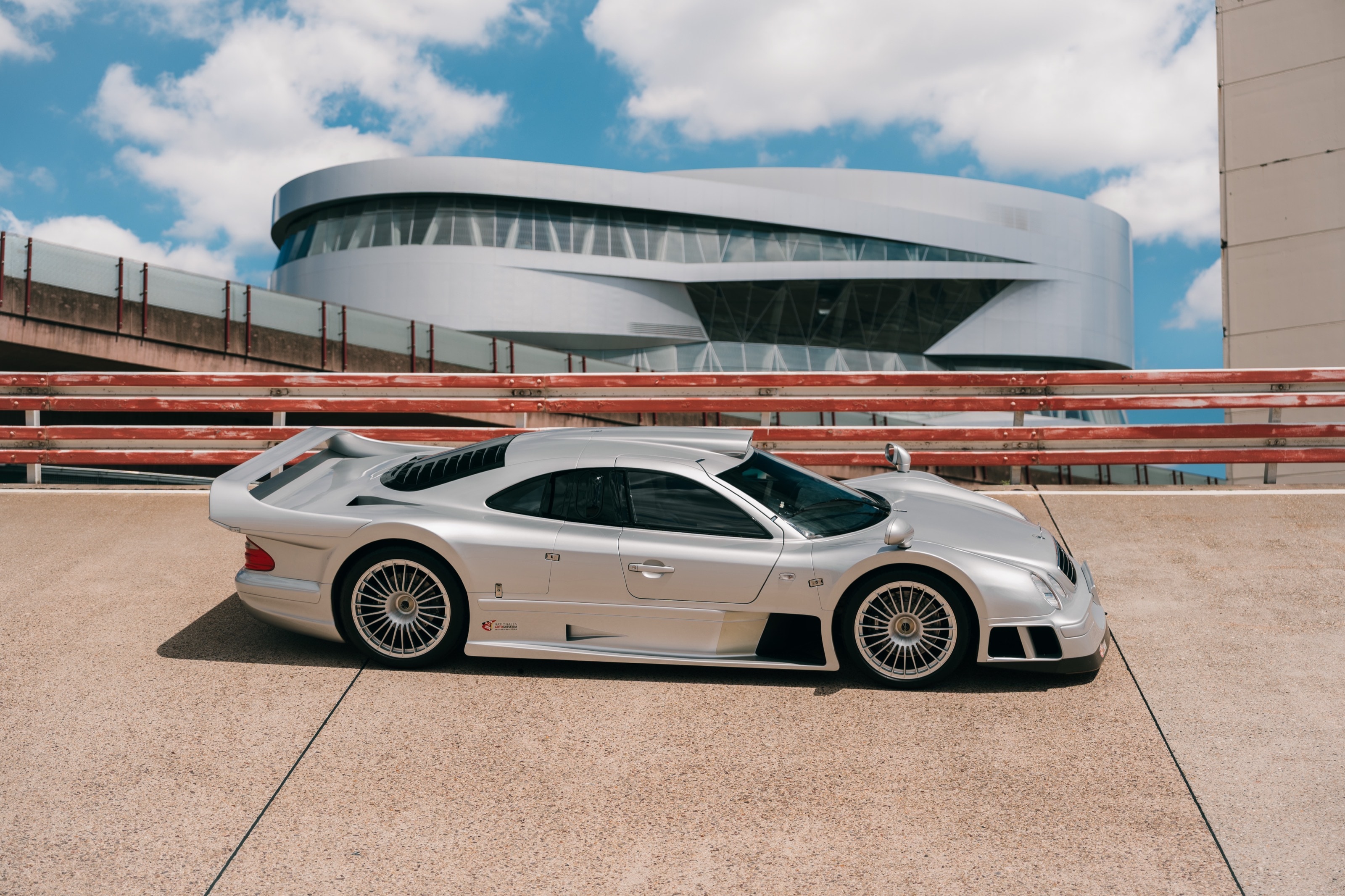 Seitenprofil des silbernen CLK GTR auf der Einfahrbahn, dahinter die Rundfassade des Mercedes-Benz-Museums