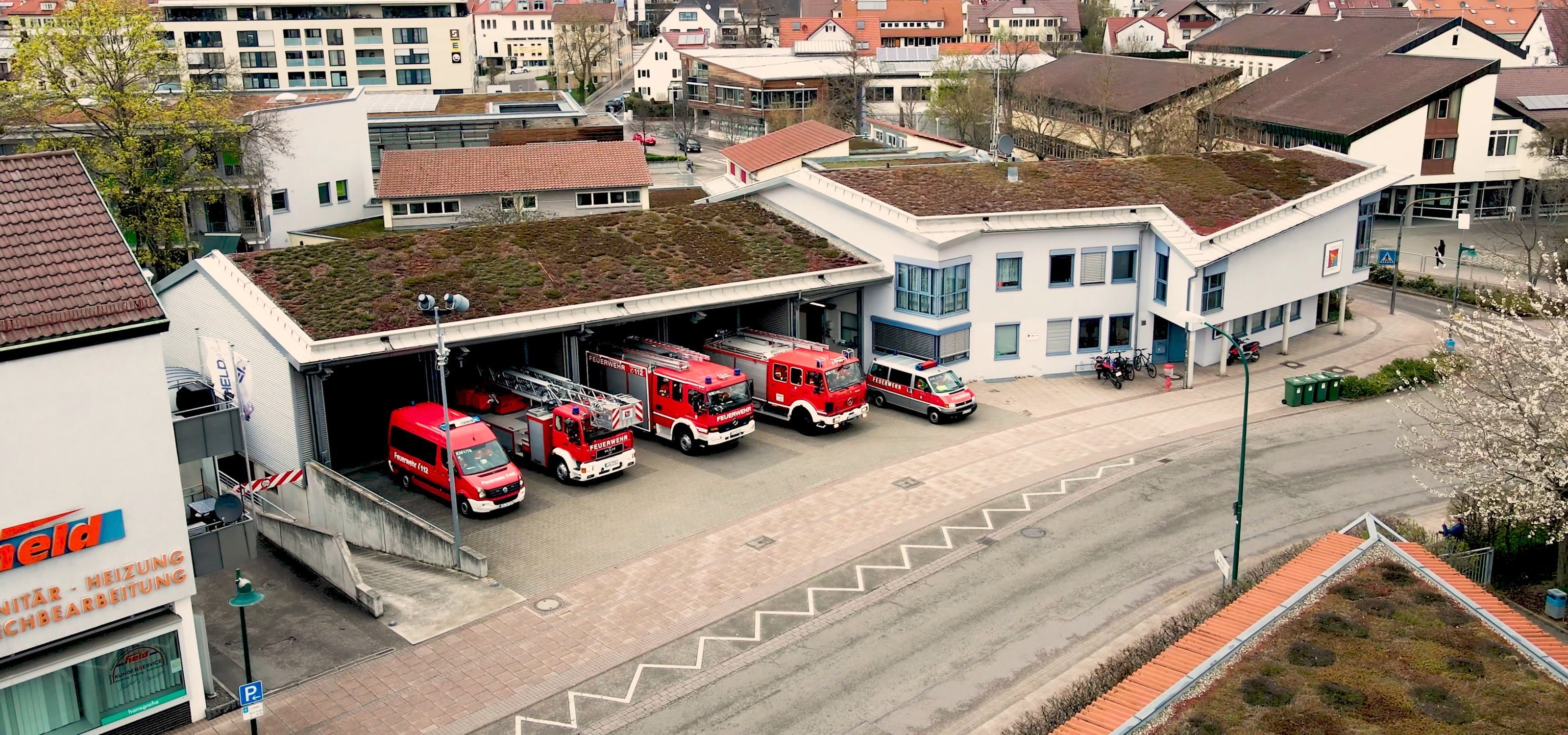 Luftbild der Feuerwache mit begrüntem Dach, in der offenen Fahrzeughalle stehen fünf rote Einsatzfahrzeuge