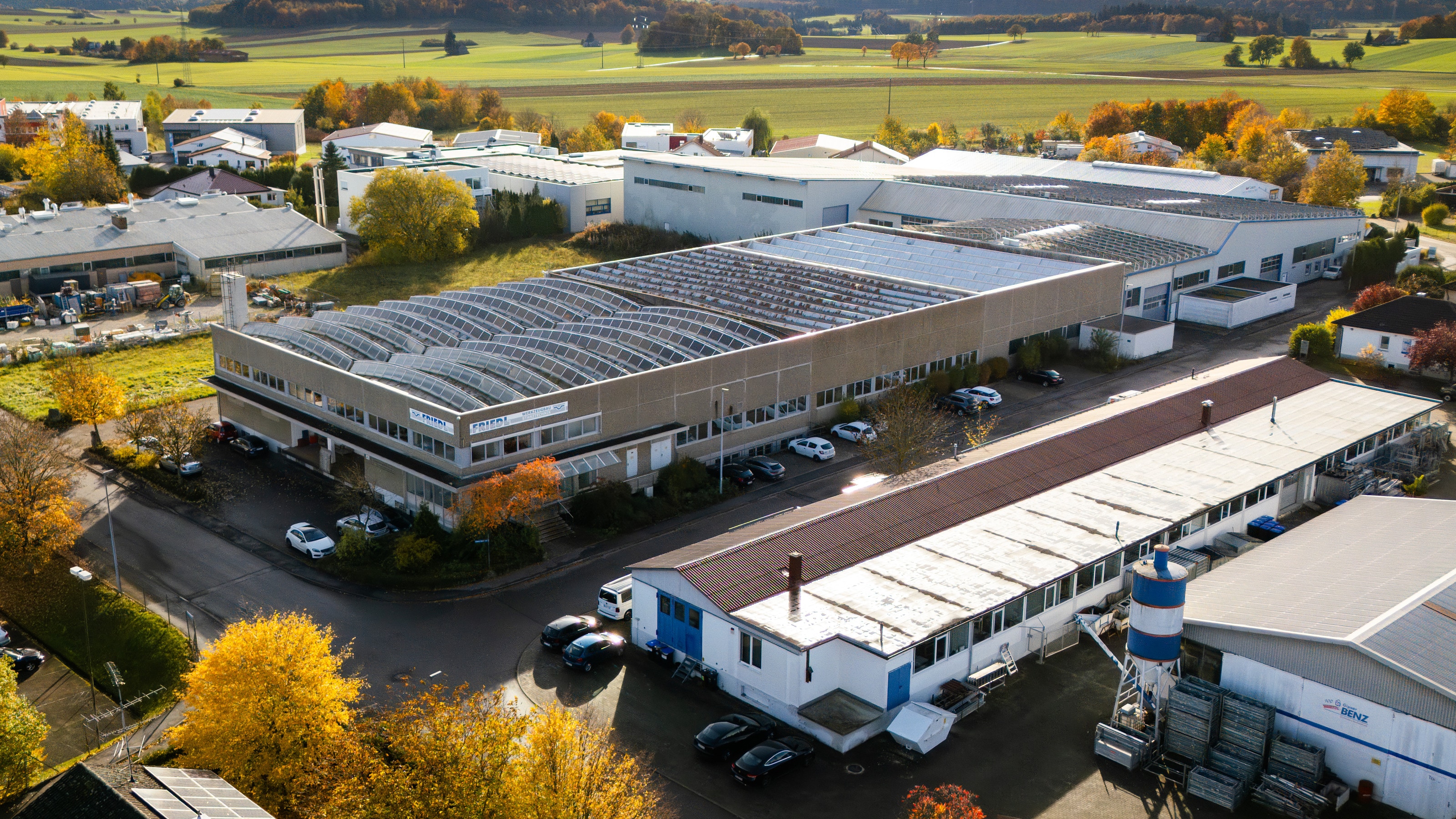 Luftbild des Firmengeländes mit Produktionshalle und Bürotrakt, dahinter Felder am Ortsrand