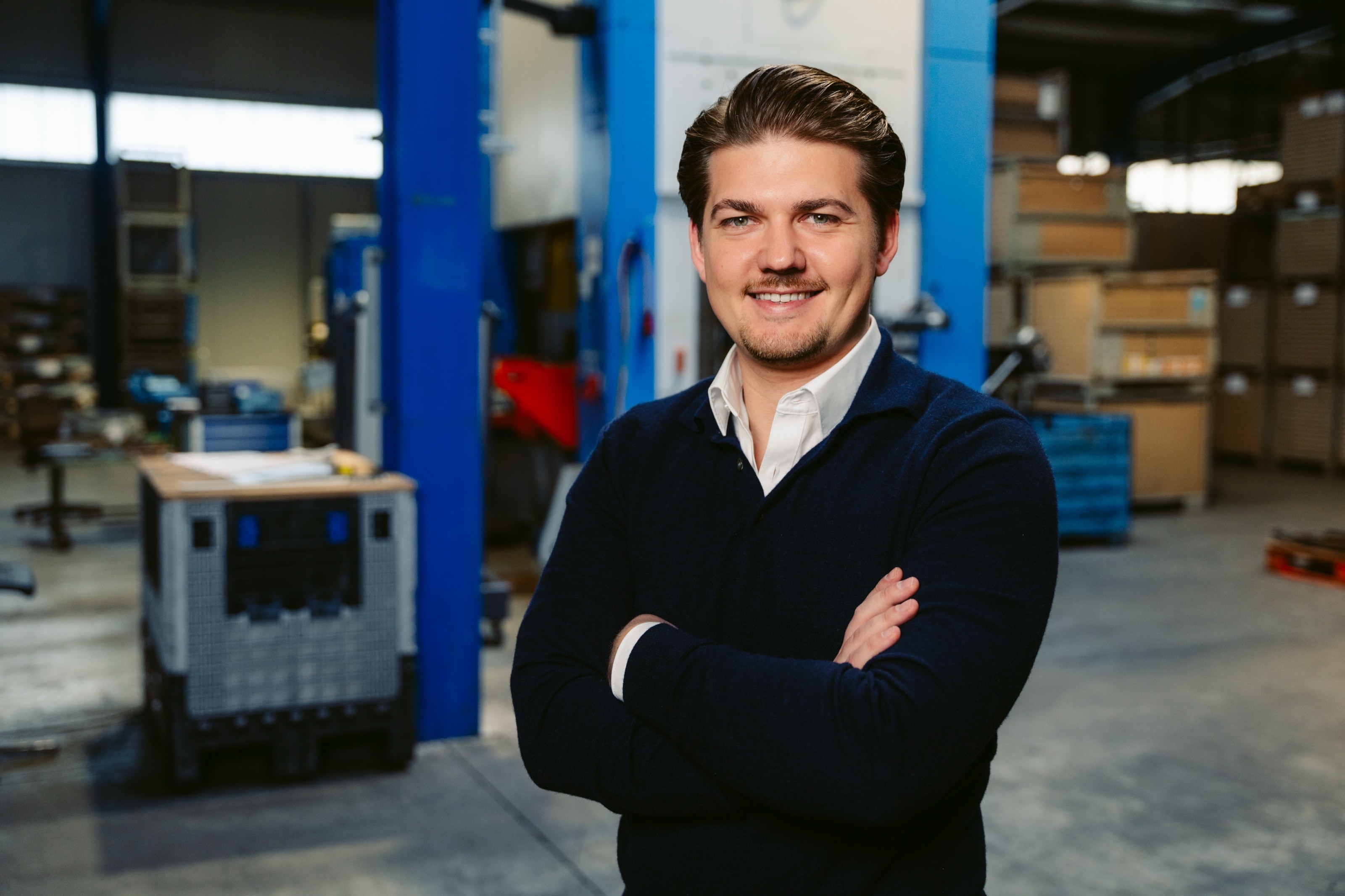 Mann im dunklen Pullover steht lächelnd mit verschränkten Armen in der Produktionshalle