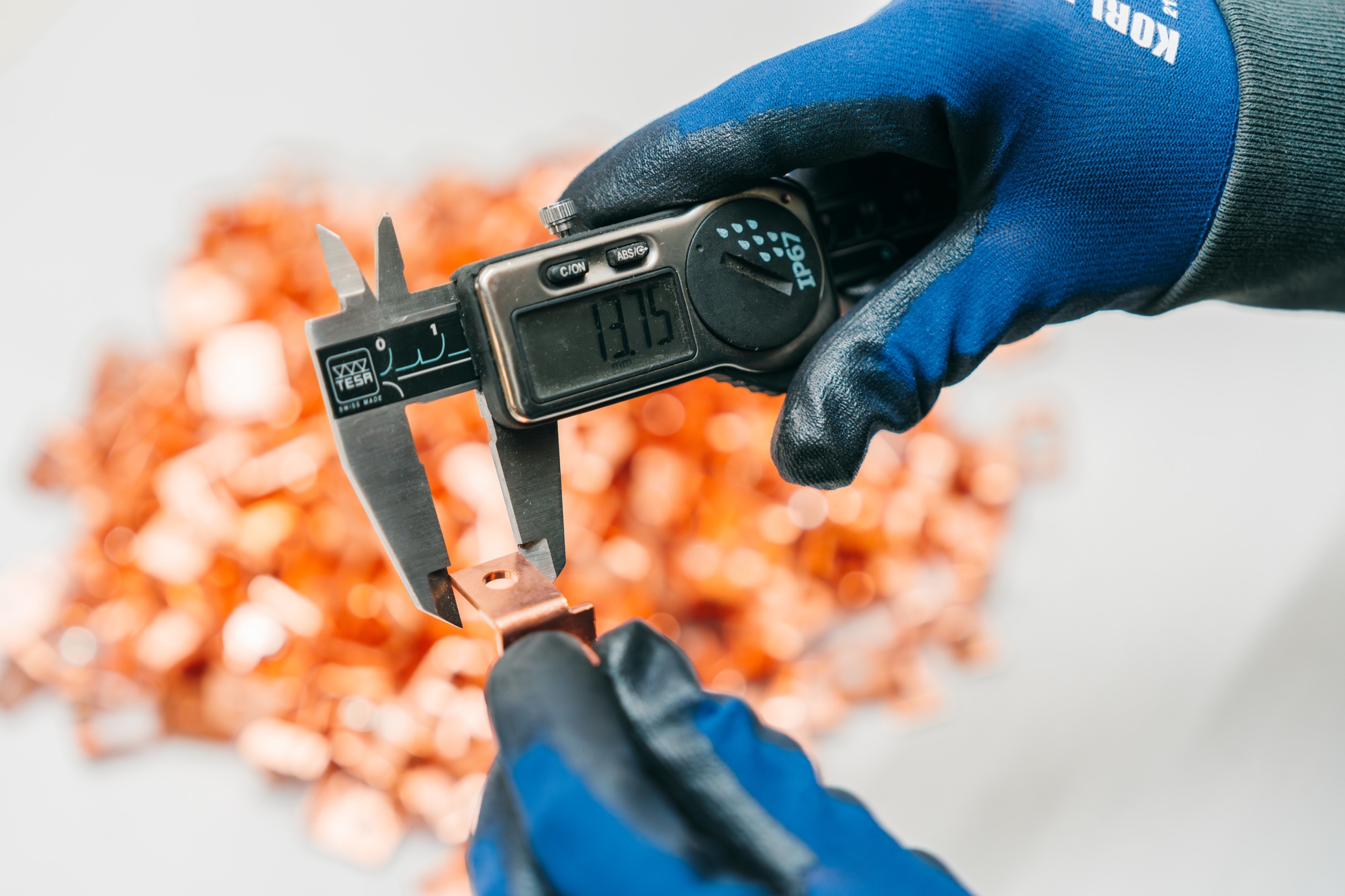 Hände in blauen Handschuhen messen ein Kupferteil mit dem digitalen Messschieber