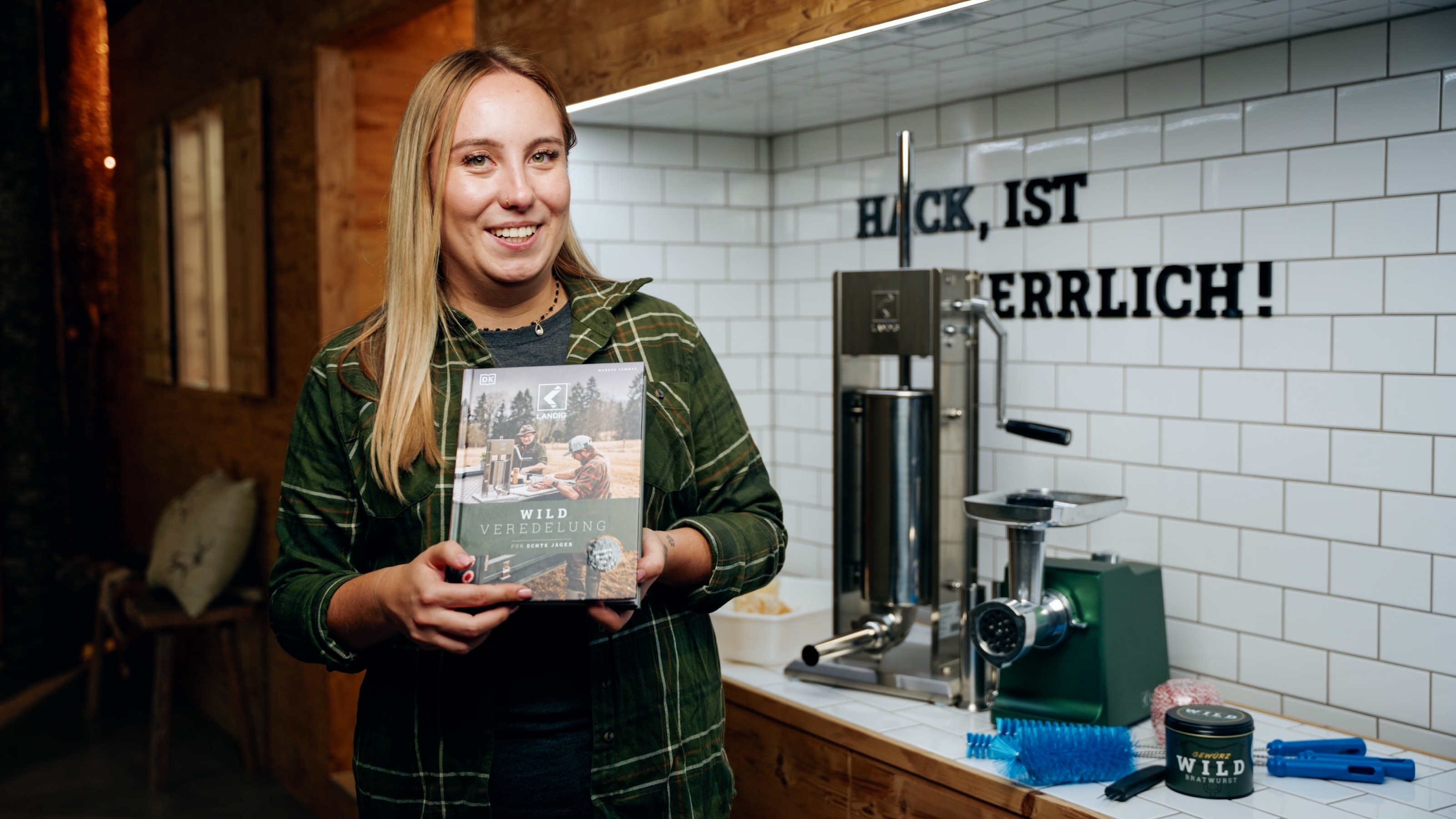 Moderatorin im grünen Karohemd hält ein Buch in die Kamera, dahinter Wurstfüller und Fleischwolf vor dem Schriftzug „Hack, ist das herrlich!“