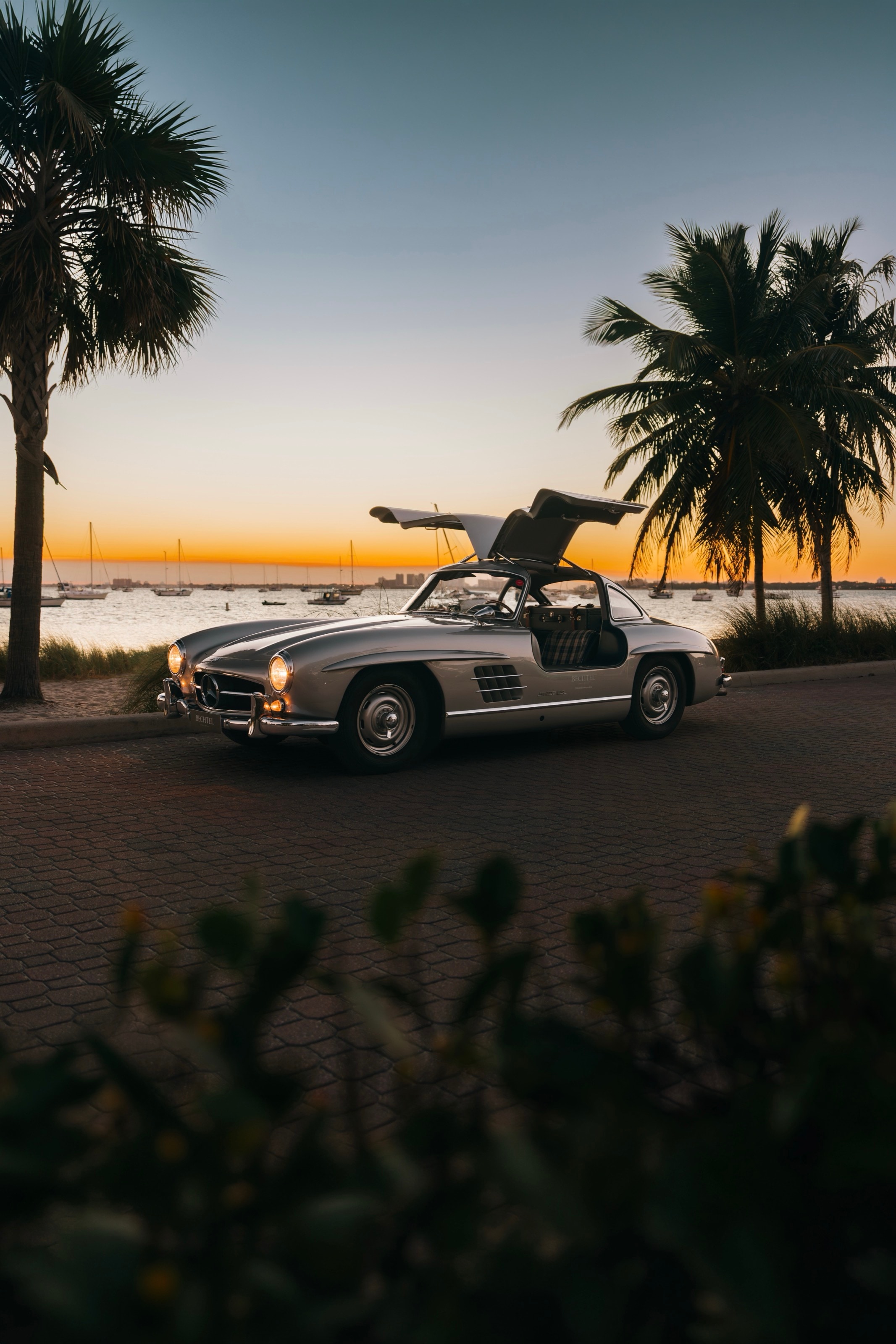 Der silberne 300 SL mit geöffneten Flügeltüren an einer Uferpromenade mit Palmen bei Sonnenuntergang