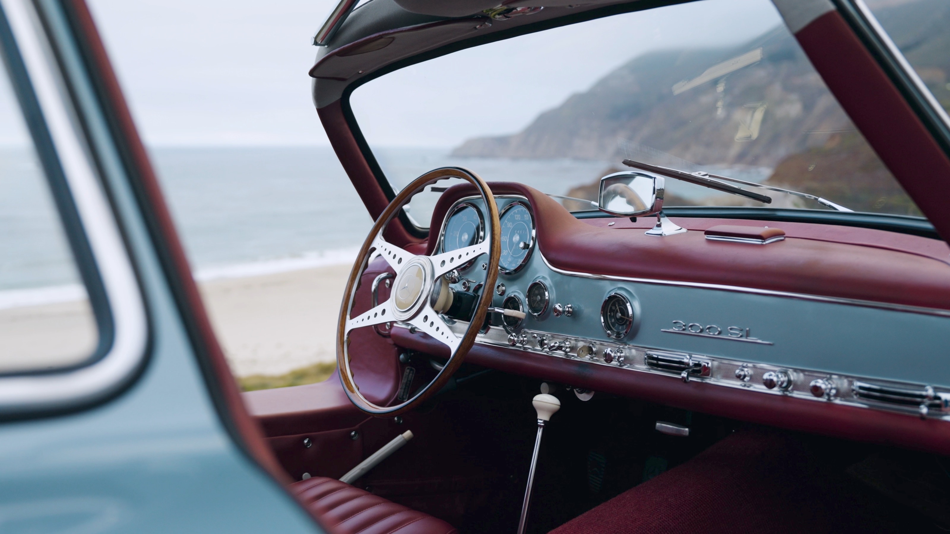 Cockpit mit rotem Leder, Holzlenkrad und 300-SL-Schriftzug, durch die geöffnete Flügeltür hindurch Strand und Meer
