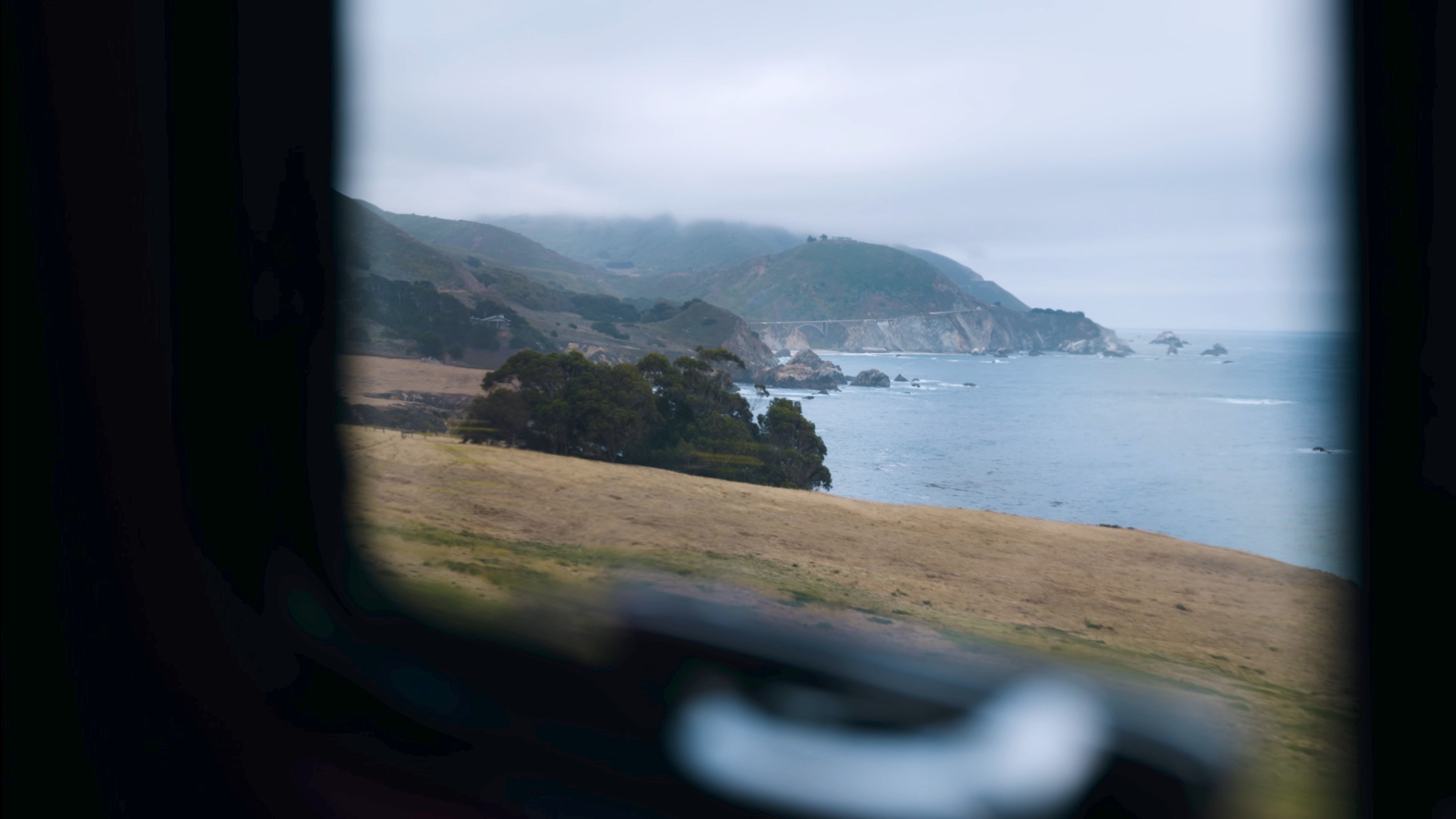 Blick durch das Seitenfenster des Gullwing auf die Steilküste von Big Sur, darüber Nebel auf den Hügeln