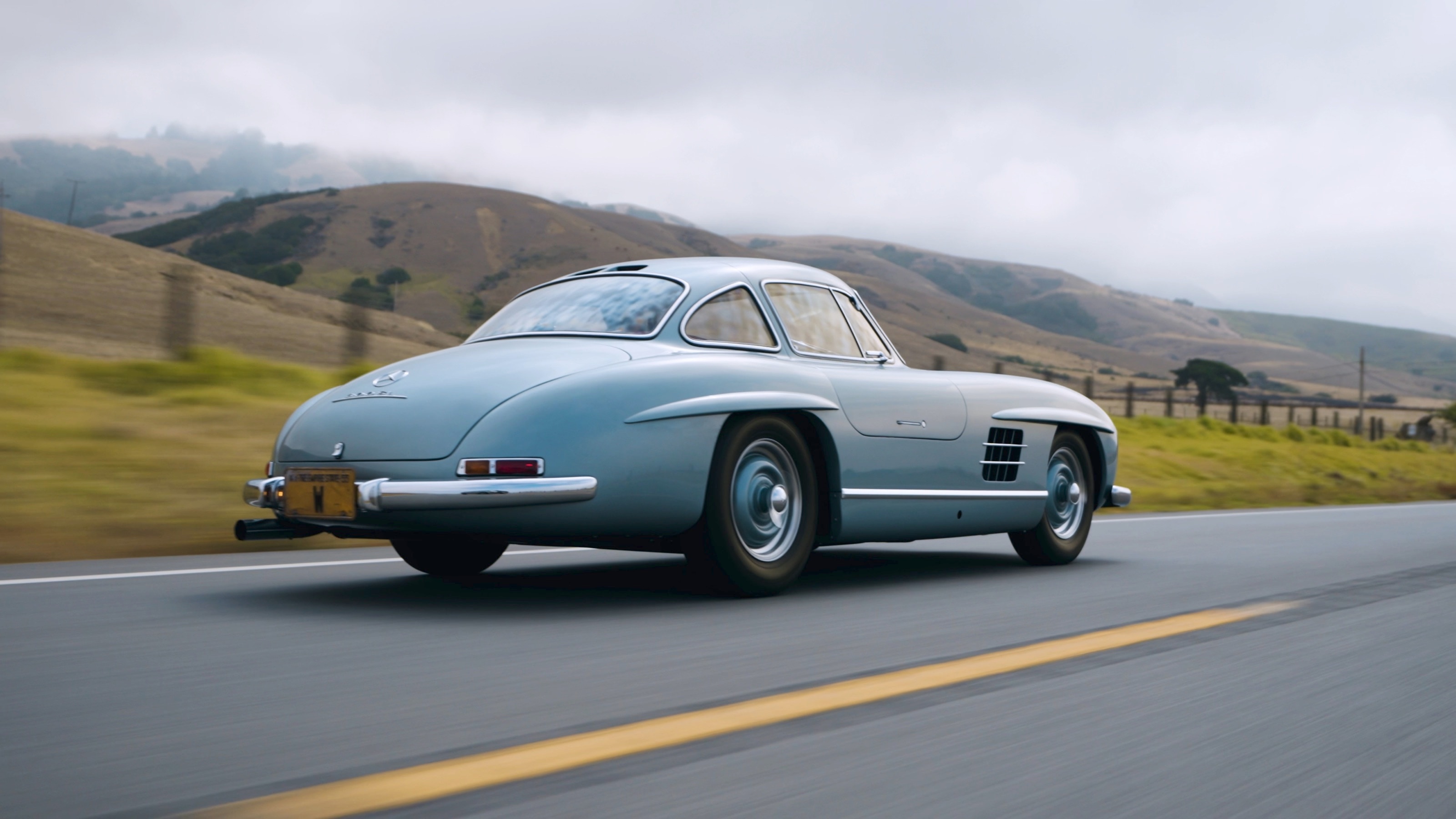 Der hellblaue 300 SL von hinten auf einer Landstraße mit gelber Mittellinie, über den Hügeln hängt Nebel