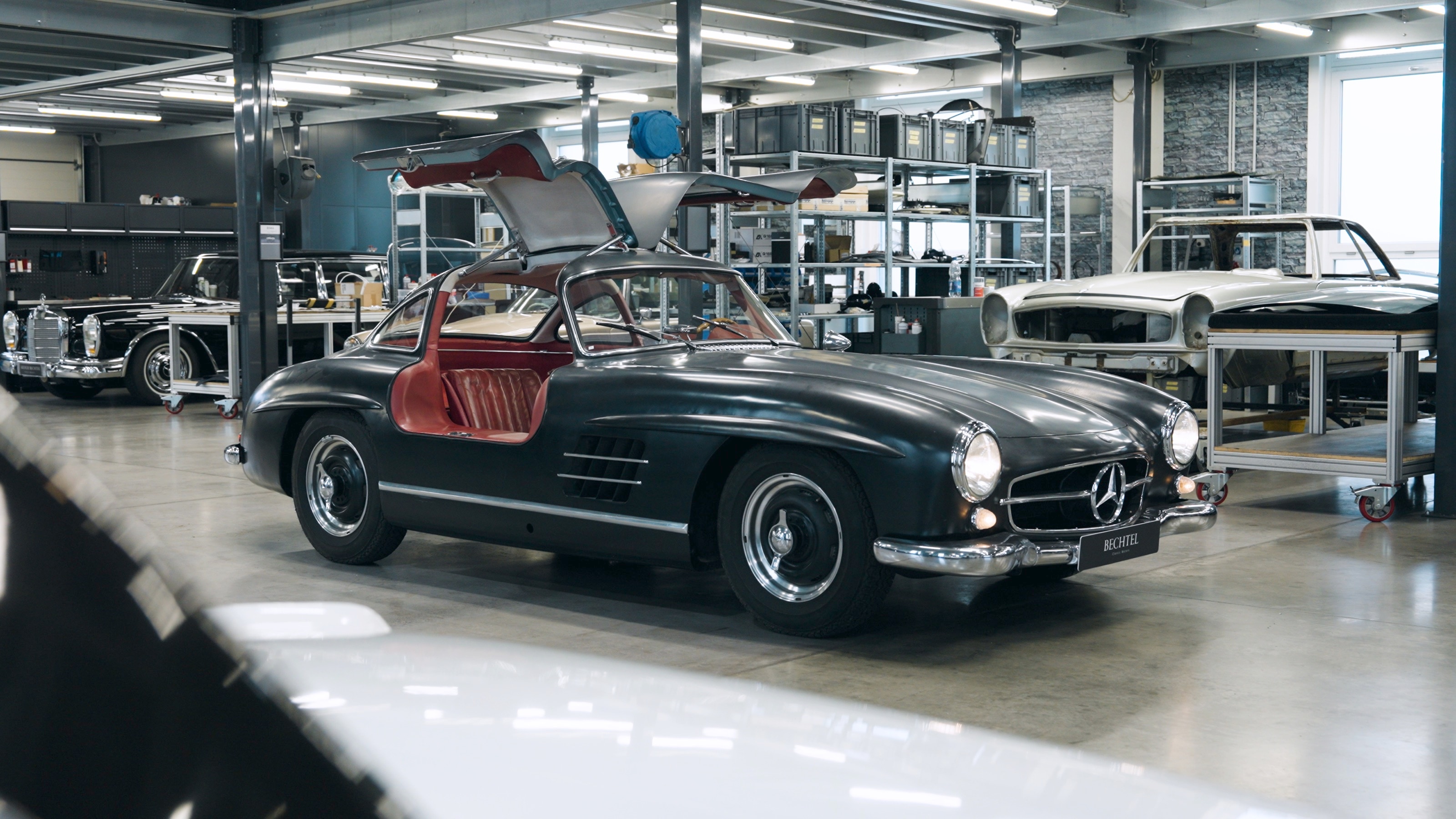 Ein anthrazitfarbener 300 SL Gullwing mit geöffneten Flügeltüren in der Werkstatt, ringsum weitere Mercedes-Benz-Klassiker
