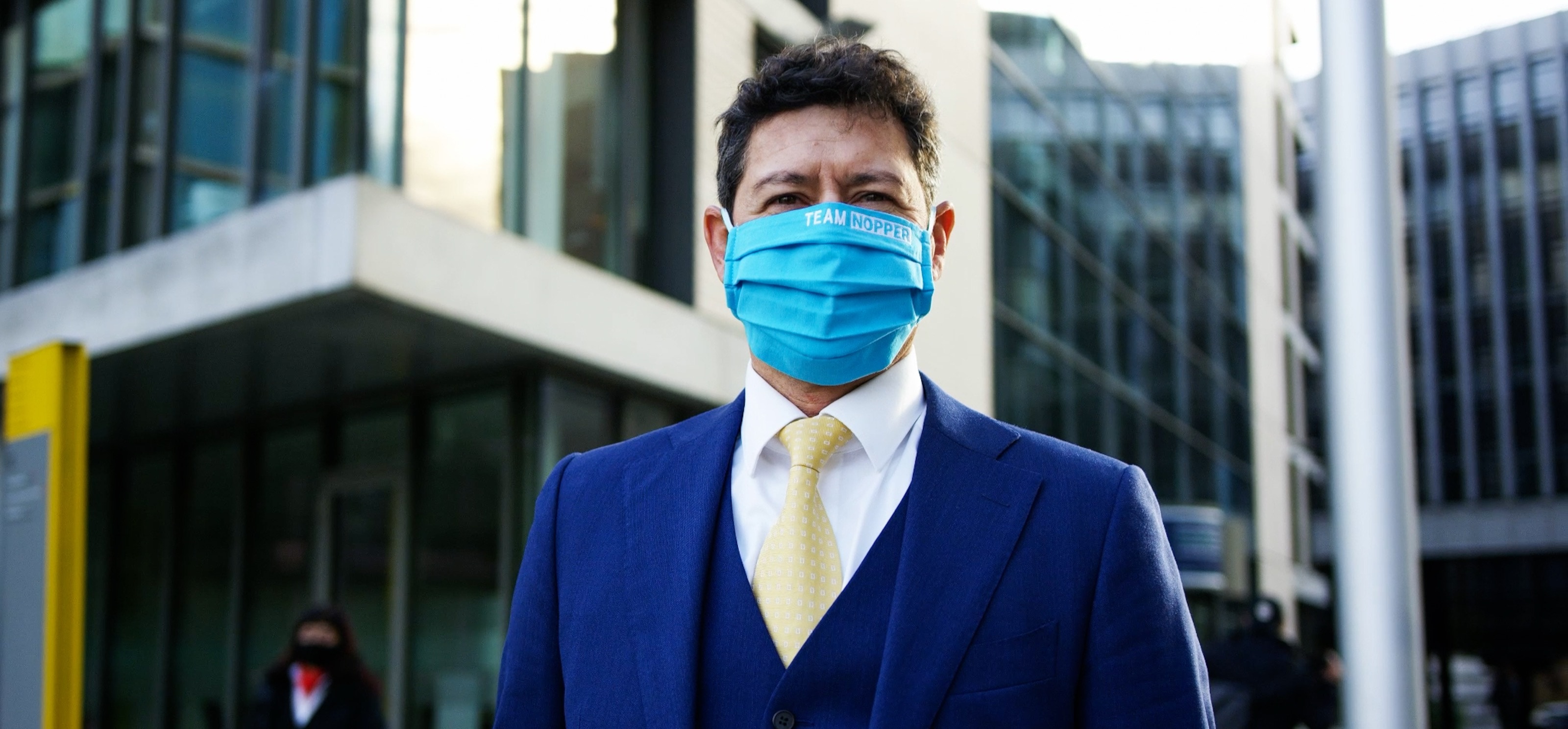 Mann im blauen Anzug mit gelber Krawatte und Mund-Nasen-Schutz vor modernen Bürogebäuden