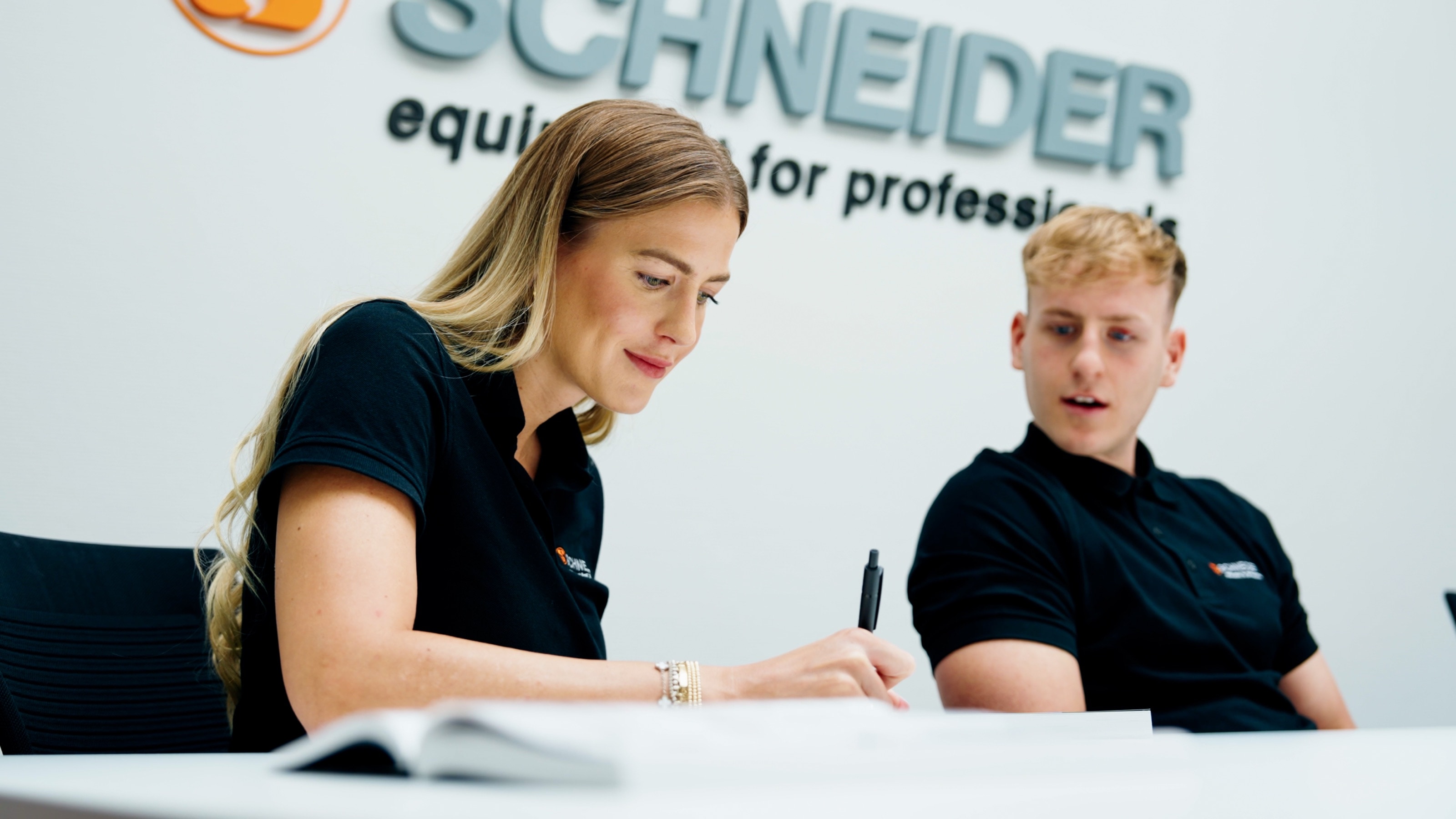 Mitarbeiterin im schwarzen Poloshirt schreibt am Besprechungstisch mit, neben ihr hört ein Kollege zu, dahinter der Schriftzug „equipment for professionals“
