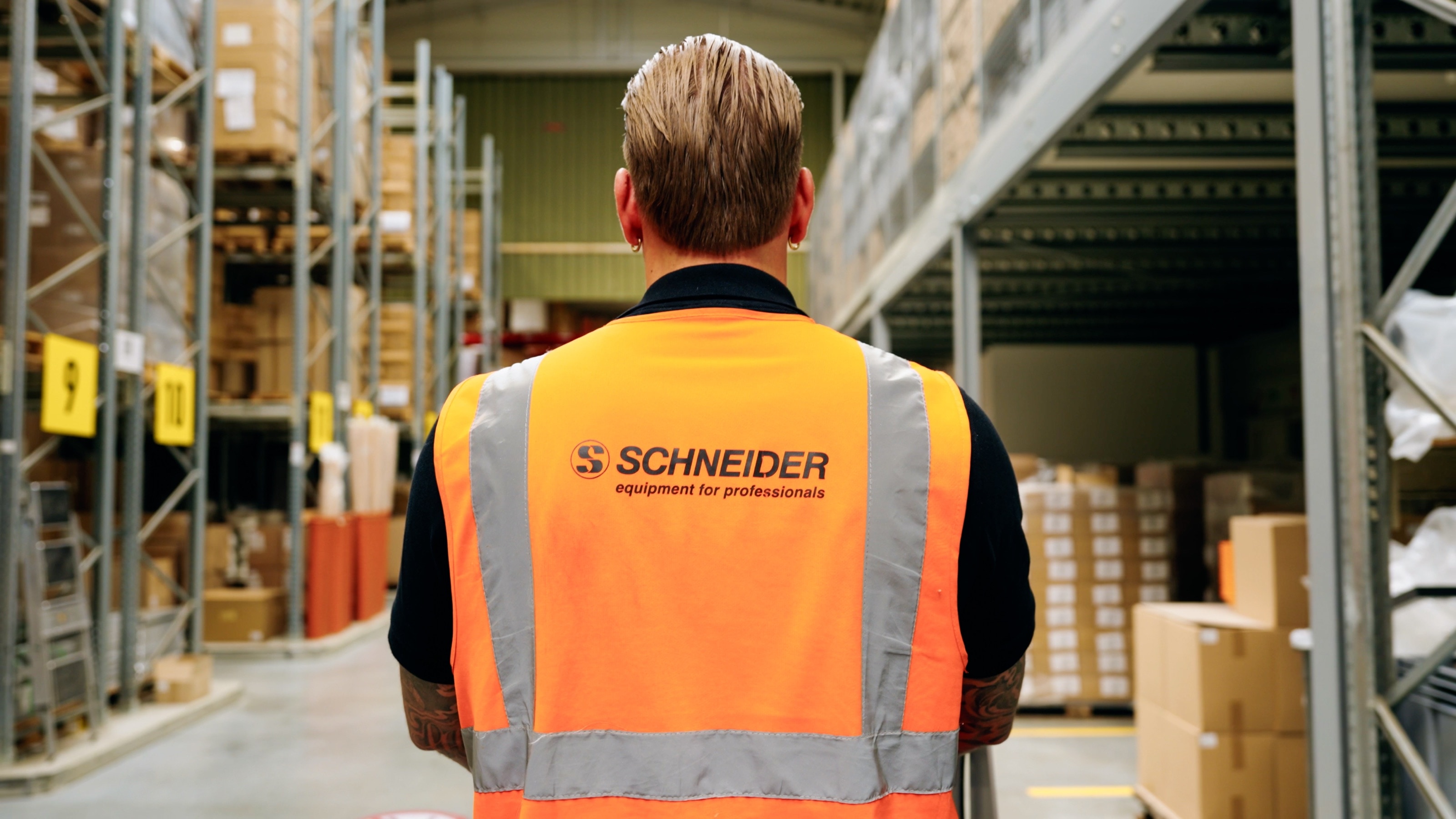 Mitarbeiter von hinten im Lager, auf seiner orangefarbenen Warnweste der Schriftzug „Schneider – equipment for professionals“
