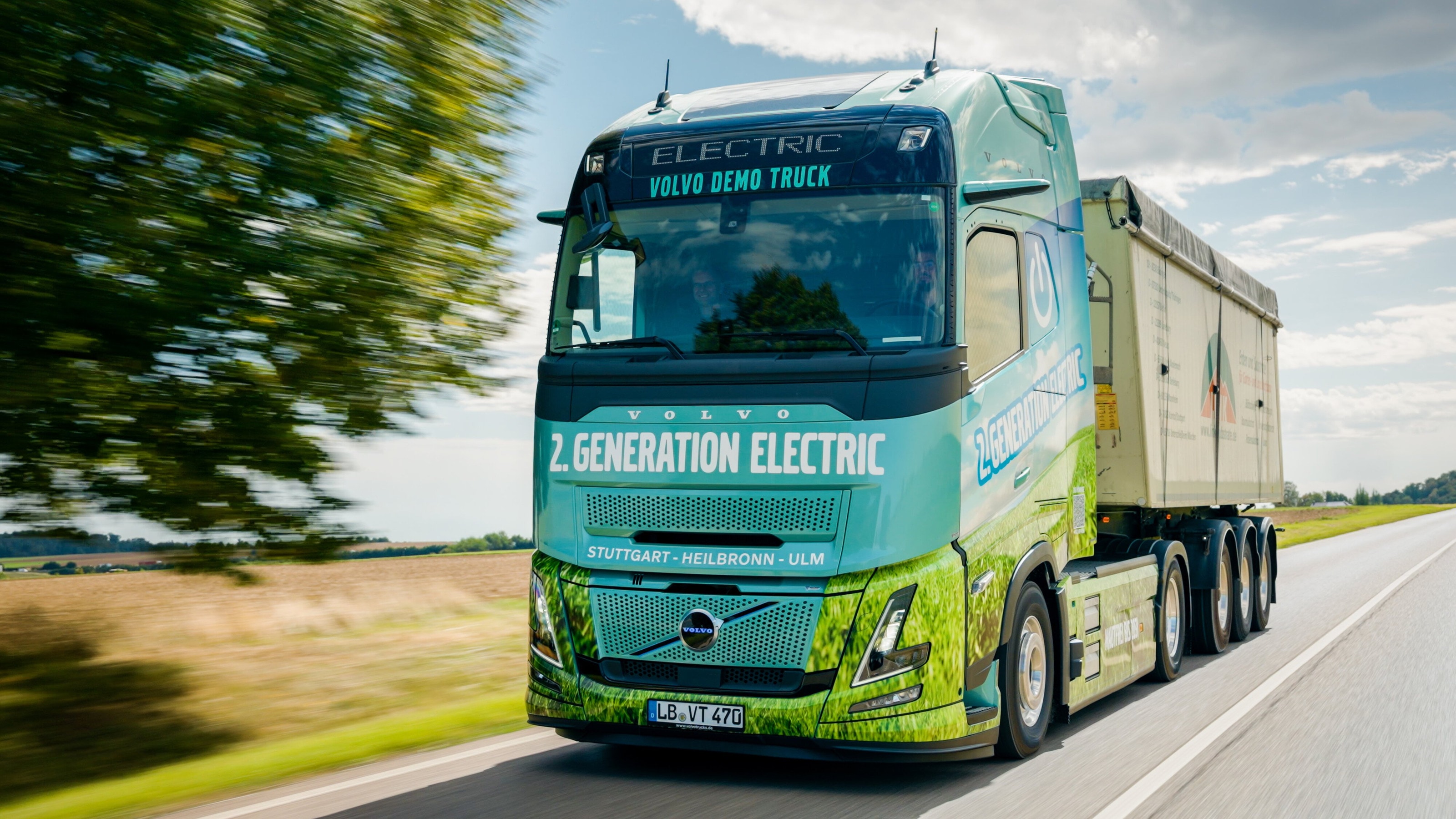Mitzieher des Volvo FH Aero Electric auf der Landstraße, im Vordergrund verwischt eine Baumreihe